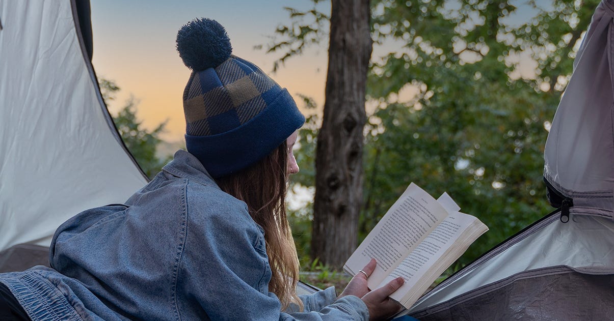 Girl sits inside a tent reading in the plaid knit watch cap with a setting sun behind her